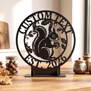 May include: A circular black metal sign featuring a cutout design of a squirrel holding an acorn surrounded by oak leaves. The text CUSTOM TEXT is arched across the top, and EST. 2026 is arched across the bottom. The sign stands on a flat base.