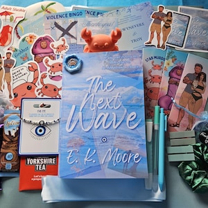 Peut inclure: Un livre intitulé The Next Wave par E.K. Moore entouré de divers articles dont des autocollants de crabes et de couples, un bracelet mauvais œil, une barre chocolatée Cadbury Dairy Milk, du thé Yorkshire, deux stylos bleu clair et un chouchou vert foncé.