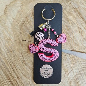Könnte beinhalten: Ein Schlüsselanhänger mit einem großen rosa Buchstaben S mit schwarzem Leopardenmuster, befestigt an einem silbernen Metallring. Das Accessoire enthält eine schwarze Quaste, eine runde Perle und zwei herzförmige Anhänger mit passendem Leopardenmuster.