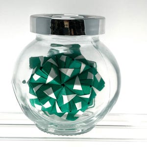 Pode incluir: Um frasco de vidro transparente e redondo com uma tampa de metal prateado brilhante. Dentro do frasco encontra-se uma escultura de papel geométrica e multifacetada com um padrão repetitivo de triângulos verde-azulados e brancos.