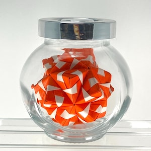 Puede incluir: Un frasco de vidrio transparente con tapa de metal plateado que contiene una escultura de origami geométrica. La escultura está hecha de papel plegado en naranja vibrante y blanco, con una forma esférica compleja y multifacética.
