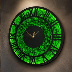 May include: A large circular wall clock featuring a black frame with Roman numerals, a central black dial with thin gold hands, and a vibrant green preserved moss ring filling the space between the center and the outer numeral border.