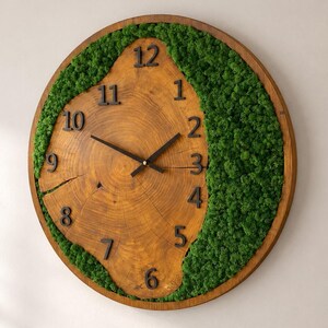 May include: A round wall clock featuring a natural wood slice face with visible grain and a crack, surrounded by a thick crescent of vibrant green preserved moss. The clock has black Arabic numerals and thin black metal hour and minute hands.