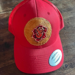 May include: A bright red baseball cap with a mesh back and a circular tan leather patch on the front. The patch features a detailed embossed and painted red rose design. A silver and black sticker on the brim reads The Authentic Yupoong Snapback.