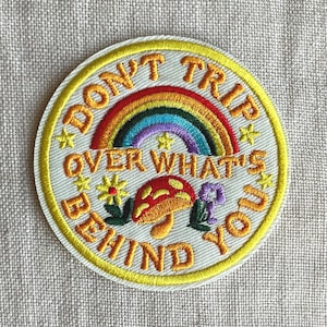 May include: A circular embroidered patch with a white background and a yellow border. It features a rainbow, a red and yellow mushroom, and small flowers. The text reads DON'T TRIP OVER WHAT'S BEHIND YOU in orange letters with yellow stars.