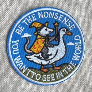 May include: A circular embroidered patch with a blue background featuring a white goose carrying an opossum wearing a yellow wizard hat and robe. The opossum holds a star-tipped wand. The text reads BE THE NONSENSE YOU WANT TO SEE IN THE WORLD.
