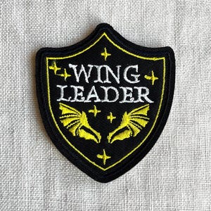 May include: A shield-shaped embroidered patch with a black background and a yellow border. The text WING LEADER is stitched in white. Below the text are two yellow wings, and the design is accented with small yellow four-pointed stars.