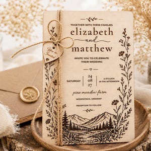 May include: A rectangular wooden wedding invitation card featuring dark brown engraved text and a mountain landscape illustration. The card is tied with twine and a small heart-shaped wooden tag reading You're Invited, framed by floral borders.