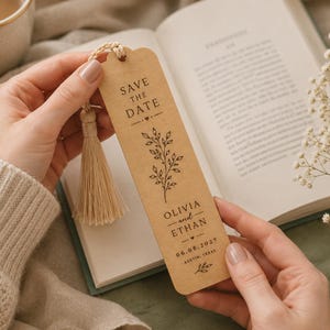 May include: A wooden bookmark with a tassel is held over an open book. The bookmark has the text "Save the Date" and the names "Olivia and Ethan" with a date and location. The bookmark has a floral design.