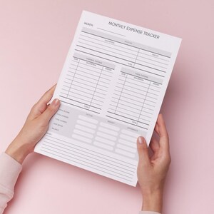 Può includere: Un tracker delle spese mensili bianco con il titolo "MONITORAGGIO DELLE SPESE MENSILI" è tenuto su uno sfondo rosa. Il tracker include sezioni per entrate, spese e dettagli del budget. Il documento è tenuto da due mani.