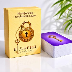 Op de afbeelding: Een gele rechthoekige kaartendoos met een illustratie van een gouden hangslot en sleutel, voorzien van de tekst "Метафоричні асоціативні карти" en "ВІДКРИЙ в собі силу". Naast de doos ligt een stapel kaarten met een paarse achtergrond en een gouden sleutel.