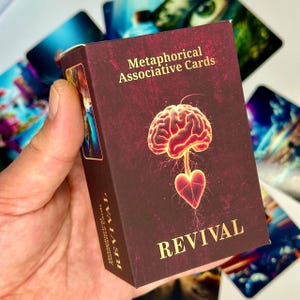 May include: A maroon card box with gold lettering reading "Metaphorical Associative Cards" and "REVIVAL". The box design features a glowing brain connected to a heart. Other cards are visible in the background.