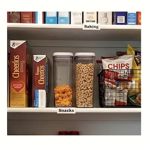 May include: A pantry shelf is organised with cereal boxes, clear containers filled with snacks, and a wire basket of crisps. The shelf is labelled "Snacks" and "Baking". The Cheerios boxes are red and yellow.