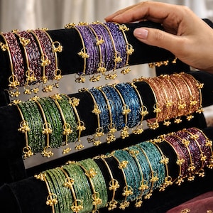 Könnte beinhalten: Eine Präsentation farbenfroher Armreifen in Lila, Blau, Grün und Orange. Jeder Armreif ist mit goldfarbenen Akzenten und kleinen, hängenden Ornamenten verziert. Die Armreifen sind auf schwarzen Samt-Präsentationsständern angeordnet.
