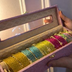 Op de afbeelding: Een rechthoekige, glitterende roze opbergdoos met een doorzichtig venster, gevuld met een collectie dunne, doorschijnende armbanden in levendig geel, turquoise, limoengroen en magenta, elk voorzien van kleine, verfijnde metalen accenten.