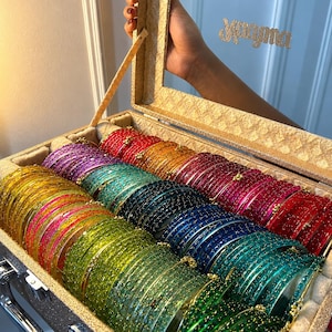 Puede incluir: Una colección de pulseras de cristal de colores en una caja decorativa. Las pulseras están dispuestas en filas, mostrando un espectro de colores que incluyen verde, azul, rojo y dorado. La caja tiene un acabado dorado brillante y un espejo.