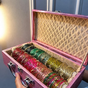 May include: A pink glittery rectangular storage case with a hinged lid containing multiple rows of thin bangles in various colors including red, green, yellow, and pink. Each bangle features small gold-toned accents and a metallic finish.