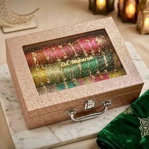 Pode incluir: Uma caixa de armazenamento retangular com glitter ouro rosa, tampa de vidro transparente e alça de metal prateado. A caixa contém fileiras de pulseiras coloridas em tons de vermelho, verde e amarelo, com o texto Eid Mubarak na tampa.