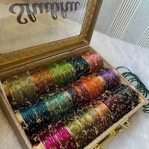 Op de afbeelding: Een verzameling veelkleurige metalen armbanden gerangschikt in een rechthoekige opbergdoos met een doorzichtig deksel. De armbanden hebben een dunne, metallic afwerking in tinten roze, groen, goud, oranje, paars, blauw en zwart met gouden kraaltjes.