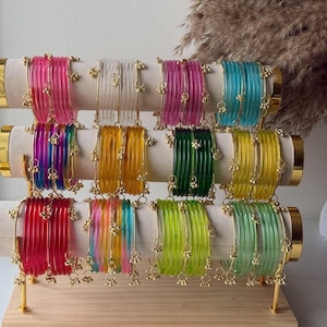 Könnte beinhalten: Eine Präsentation farbenfroher Armreifen in verschiedenen Farbtönen, darunter Rosa, Transparent, Lila und Grün. Jeder Armreif ist mit kleinen goldfarbenen Glöckchen verziert. Die Armreifen sind auf einem Holzständer mit beigen zylindrischen Stützen angeordnet.