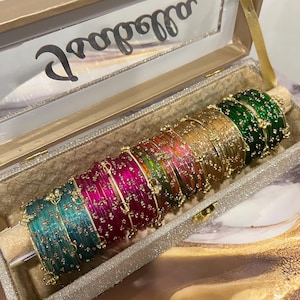 Könnte beinhalten: Ein Set aus mehrfarbigen, durchscheinenden Armreifen mit kleinen goldfarbenen Blumenakzenten, die in einer Reihe in einer rechteckigen, beigen, glitzernden Aufbewahrungsbox mit transparentem Deckel und dem Schriftzug "allodose" liegen.
