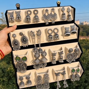 Könnte beinhalten: Ein schwarzer Schmuckkoffer mit einer Sammlung silberfarbener Ohrringe. Das Sortiment umfasst Ohrringe im Jhumka-Stil, Schmetterlingsmotive, kreisförmige Designs und von Pfauen inspirierte Stücke mit grünen Akzenten, die alle in Reihen angeordnet sind.
