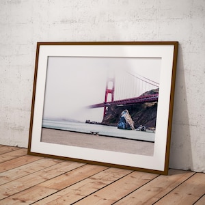 Può includere: Una fotografia paesaggistica incorniciata che ritrae il Golden Gate Bridge parzialmente oscurato da una fitta nebbia bianca. La struttura rossa del ponte si estende su acque scure e scogliere rocciose, con una riva sabbiosa in primo piano.