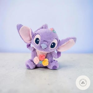 May include: A plush, lavender-colored character with large ears and big eyes, adorned with a floral arrangement. The stuffed animal has a light blue nose and pink inner ears. The toy is a popular character from a well-known animated film.