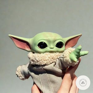 Puede incluir: Mu&ntilde;eco de piel verde con grandes ojos negros y orejas puntiagudas, que lleva un abrigo beige con cuello esponjoso. La mano derecha del mu&ntilde;eco est&aacute; extendida. El mu&ntilde;eco es un personaje de una popular serie de ciencia ficci&oacute;n.