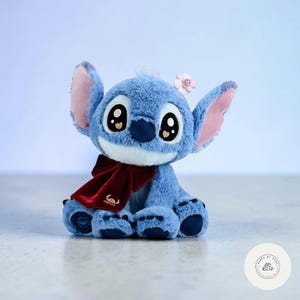May include: A plush blue character with large eyes, pink-lined ears, and a small flower on its head. It wears a burgundy bow and has a white muzzle. The plush toy is a popular character from a Disney animated film.