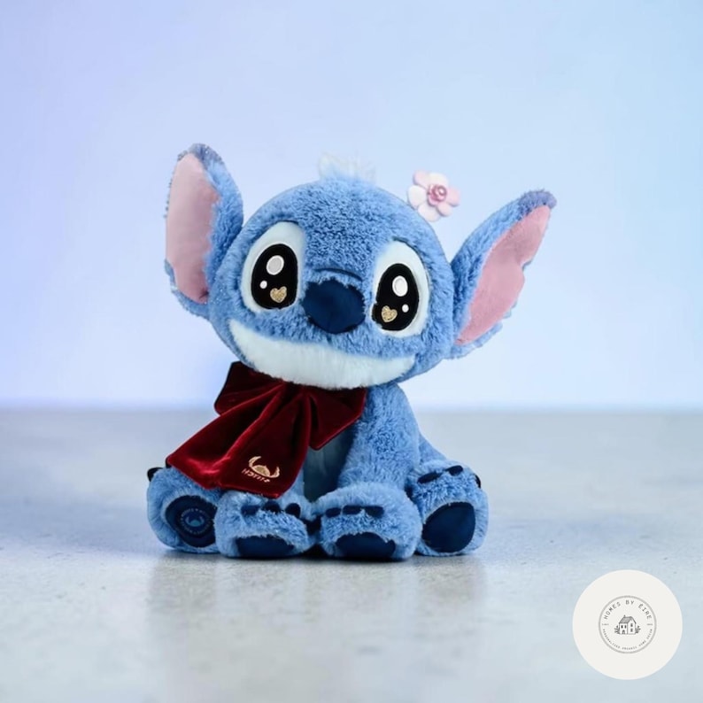 May include: A plush blue character with large eyes, pink-lined ears, and a small flower on its head. It wears a burgundy bow and has a white muzzle. The plush toy is a popular character from a Disney animated film.