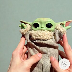 Puede incluir: Mu&ntilde;eco de personaje de piel verde con ojos y orejas grandes, que lleva una t&uacute;nica color canela con un cuello difuso. El mu&ntilde;eco est&aacute; sostenido por dos manos. El mu&ntilde;eco es un personaje de la franquicia Star Wars.