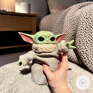 Puede incluir: Una figura verde de un personaje con grandes ojos negros y orejas puntiagudas, que lleva una t&uacute;nica beige con cuello y pu&ntilde;os esponjosos. La figura se sostiene en una mano, con un fondo borroso de una cama y una mesita de noche.