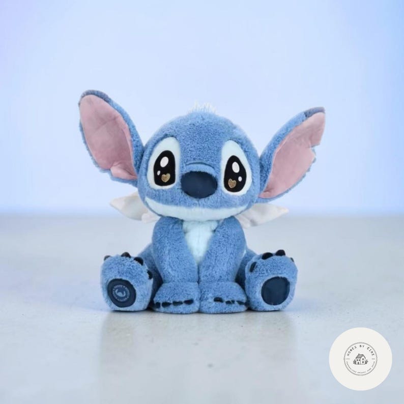 May include: A plush blue Stitch doll with large ears, pink inner ears, and big black eyes. The doll has a white chest and small white wings. The doll is sitting with its legs outstretched and its arms at its sides.