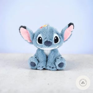 May include: A plush toy of a blue alien character with large ears, black eyes, and a white belly. The toy has pink inner ears and a small patch of yellow and orange on its head. It is sitting on a light-colored surface.