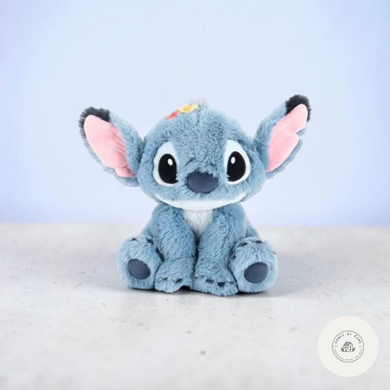 May include: A plush toy of a blue alien character with large ears, black eyes, and a white belly. The toy has pink inner ears and a small patch of yellow and orange on its head. It is sitting on a light-colored surface.