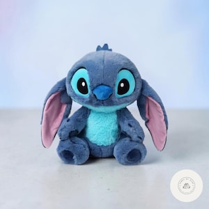 May include: A plush Stitch doll, featuring a blue body, large black eyes, and oversized ears with pink inner details. The doll has a turquoise belly and a small tuft of hair on its head. It is a soft, cuddly toy.