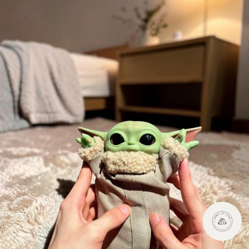 Puede incluir: Una figura verde de Grogu con grandes ojos negros y orejas puntiagudas. La figura lleva una t&uacute;nica color canela con un cuello difuso. La figura est&aacute; sujeta por dos manos. Es un objeto de colecci&oacute;n.