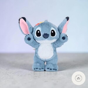 May include: A plush, blue Stitch character with large black eyes, pink inner ears, and a white belly. The toy has its arms raised and a small rainbow detail on its head. The character is standing on a light-colored surface.