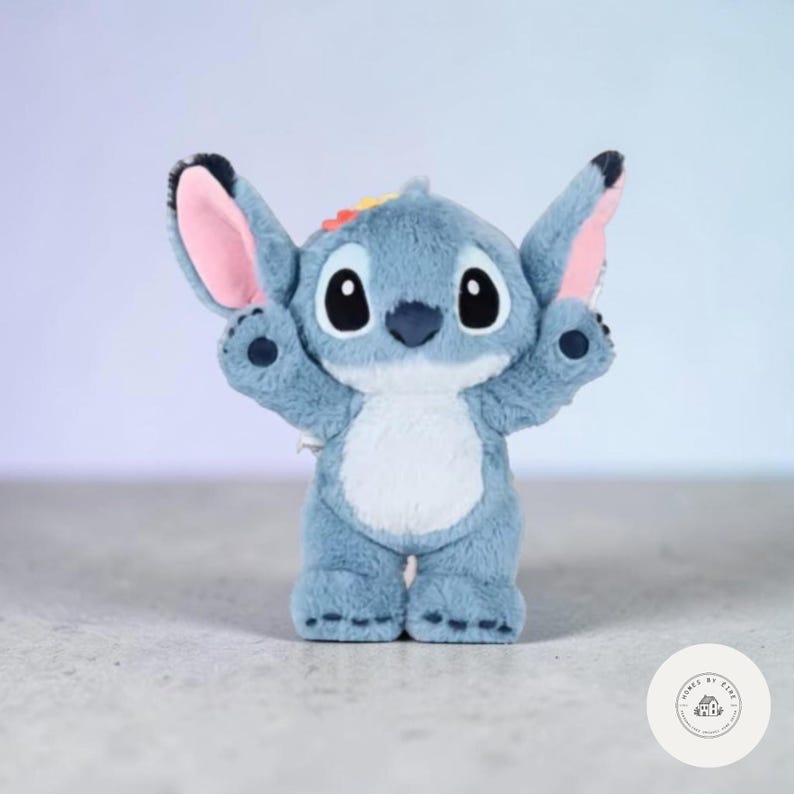 May include: A plush, blue Stitch character with large black eyes, pink inner ears, and a white belly. The toy has its arms raised and a small rainbow detail on its head. The character is standing on a light-colored surface.