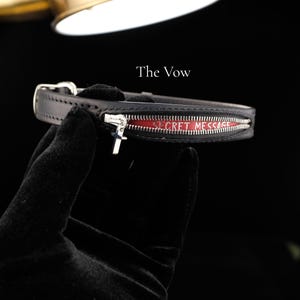May include: A black leather collar featuring a silver zipper that opens to reveal a red interior with the text SECRET MESSAGE printed in white. A small silver cross charm hangs from the zipper pull. The text The Vow appears above the collar.
