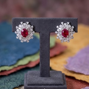 Pode incluir: Um par de brincos com um rubi vermelho central de corte oval, rodeado por um halo de diamantes brancos de corte baguette e redondo, montados em metal prateado. Os brincos estão expostos num suporte de joalharia em forma de T cinzento.