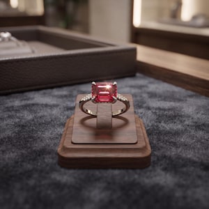 Puede incluir: Un anillo de tono plateado con una gran piedra preciosa roja rectangular de corte esmeralda en un engaste de cuatro garras. La banda está decorada con pequeñas piedras claras de corte redondo incrustadas a ambos lados del engaste.