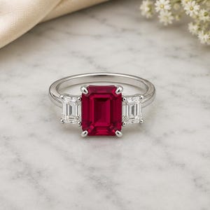 May include: A silver-toned ring featuring a large, rectangular emerald-cut red gemstone at the centre, flanked by two smaller, rectangular emerald-cut clear white gemstones on either side, all set in a polished metal band with four-prong settings.