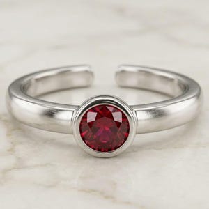 Puede incluir: Un anillo de banda abierta en tono plateado con una piedra preciosa redonda y facetada de color rojo intenso, engastada en un bisel circular liso. La banda tiene un acabado metálico pulido y una abertura en la parte posterior para ajustar.