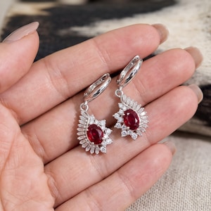 Puede incluir: Un par de pendientes colgantes de tono plateado con una piedra preciosa central de color rojo intenso en talla ovalada, rodeada por un halo de pequeñas piedras transparentes en talla baguette y redonda. Cierre de bisagra y forma de gota.