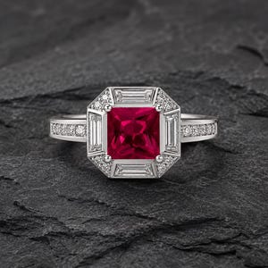 Puede incluir: Un anillo de tono plateado con una piedra preciosa de color rojo intenso de corte cuadrado en el centro. La piedra está rodeada por un halo geométrico de cuatro diamantes de corte baguette y pequeños diamantes redondos, con diamantes en la banda.