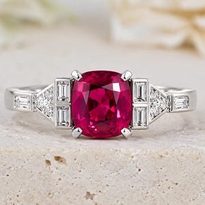 Puede incluir: Un anillo de tono plateado con una gran piedra preciosa de color rojo intenso en talla cojín en el centro. La banda está decorada con piedras rectangulares en talla baguette y pequeñas piedras redondas transparentes en patrones triangulares.