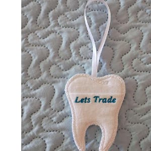 May include: A white felt tooth-shaped ornament featuring a white ribbon loop at the top for hanging. The text "Lets Trade" is embroidered in teal thread across the center of the tooth, which has a distinct two-rooted base design.