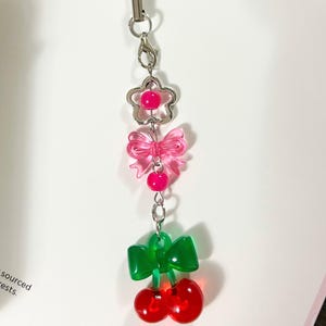 May include: A phone charm featuring a black cord, a silver-tone lobster clasp, a pink flower bead with a central pink sphere, a translucent pink bow bead, a small pink sphere, and a green bow bead attached to two translucent red cherry beads.